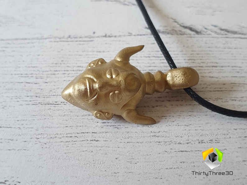 May include: Gold-colored pendant necklace featuring a detailed, stylized face with horns. The pendant is attached to a black cord.