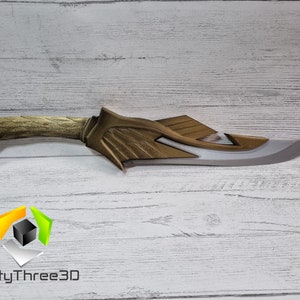 May include: A gold and silver fantasy-style dagger with a curved blade and a detailed handle. The dagger is on a white wooden surface.