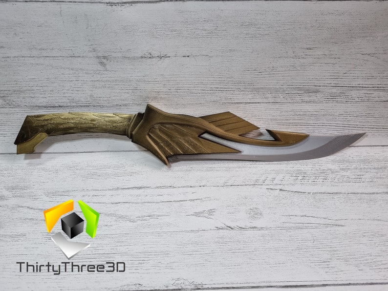 May include: A gold and silver fantasy-style dagger with a curved blade and a detailed handle. The dagger is on a white wooden surface.