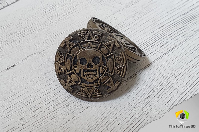May include: Two gold-colored metal coins with a skull and crossbones design. The coins are stacked on top of each other.