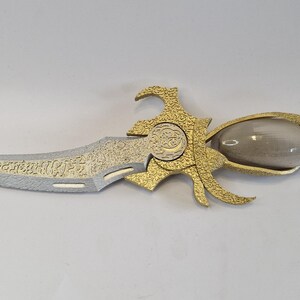 May include: An ornate dagger with a silver blade and gold-colored handle. The blade has a detailed pattern, and the handle features a bulbous, clear section and a red end. The design is intricate and decorative.