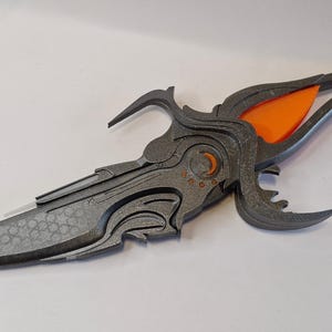 May include: A decorative dagger with a grey blade and handle, featuring intricate, stylised designs. The handle incorporates an orange accent. The blade has a textured pattern. The overall design suggests a fantasy or cosplay theme.