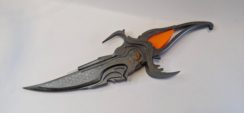 May include: A decorative dagger with a grey blade and handle, featuring intricate, stylised designs. The handle incorporates an orange accent. The blade has a textured pattern. The overall design suggests a fantasy or cosplay theme.