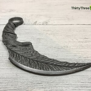 May include: A silver-colored metal throwing knife with a curved blade and a detailed feather design. The knife has a ring at the top for holding.