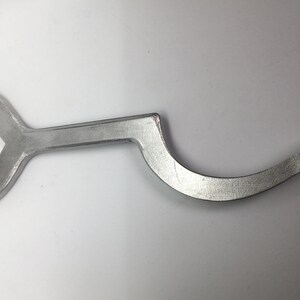Puede incluir: Una llave inglesa plateada con un mango curvo. La llave tiene una abertura en forma de U en un extremo y un extremo recto en el otro.