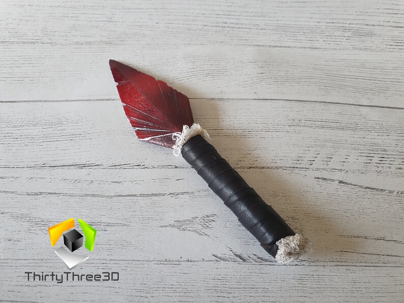 May include: A handmade decorative knife with a red blade and a black wrapped handle. The blade has a feather-like design with white accents. The handle is wrapped in black material and has a white trim at the base of the blade.