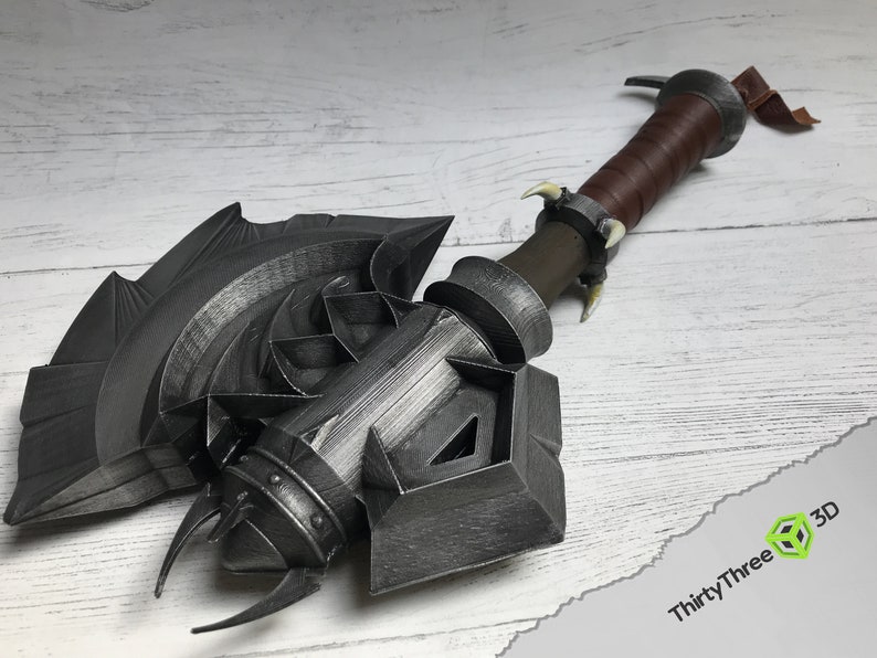 May include: A large, detailed, silver-colored axe with a leather-wrapped handle. The axe head has a complex design with sharp points and a textured surface. The handle has a brown leather wrap and a metal ring at the top.