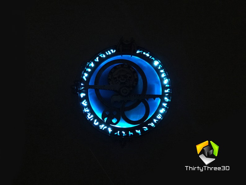 May include: A circular, black metal pendant with a blue light shining through the center. The pendant features a design of interlocking circles and runes around the edge.