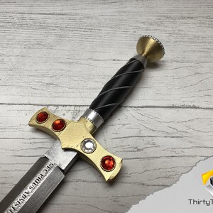 May include: A gold and silver sword with a black handle and red gems. The sword has a inscription that reads "SEC FIRIS VISII".