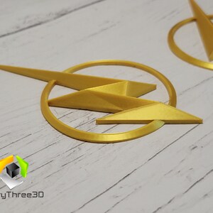 May include: Two gold-colored 3D printed lightning bolt logos. The logos are circular with a lightning bolt in the center. The logos are on a white wooden surface.