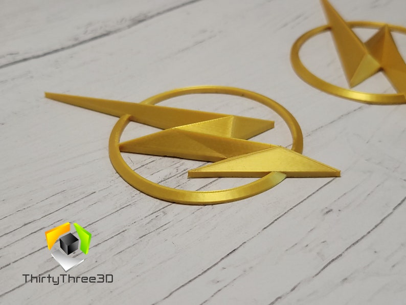 May include: Two gold-colored 3D printed lightning bolt logos. The logos are circular with a lightning bolt in the center. The logos are on a white wooden surface.
