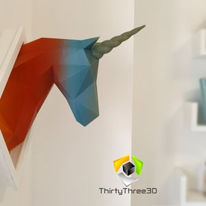 May include: A 3D printed unicorn head sculpture in a white frame. The unicorn is made of paper and is painted in shades of red, blue, and green. The sculpture is mounted on a white wall.