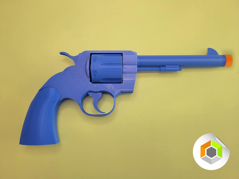 Op de afbeelding: Een blauwe speelgoedrevolver met een oranje punt, tegen een gele achtergrond. Het speelgoedpistool is een gedetailleerde replica, met een trekker, hamer en cilinder. Het algehele ontwerp is een klassieke westernstijl.