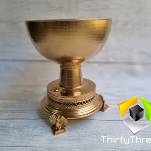 May include: A gold-colored metal cup with a decorative base and three legs. The cup is on a light wood surface.