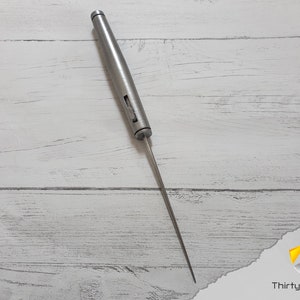 Puede incluir: Una aguja de tejer de metal plateado con una punta puntiaguda y un mango con un peque&ntilde;o agujero. La aguja est&aacute; sobre una superficie de madera blanca.