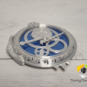 Puede incluir: Una esfera de reloj impresa en 3D de color plateado con engranajes azules y un anillo exterior plateado con marcas. La esfera del reloj est&aacute; sobre una superficie de madera blanca.