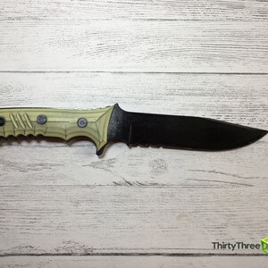 May include: A black and tan tactical knife with a serrated blade and a textured handle. The knife is lying on a white wooden surface.