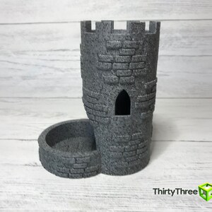 May include: A gray stone tower-shaped dice holder with a separate tray for dice. The tower has a small window opening and a textured surface.