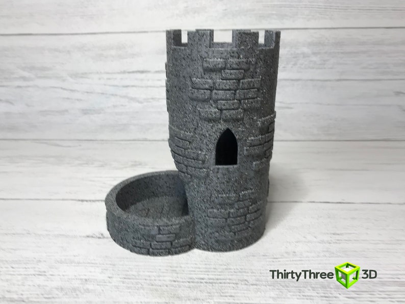 May include: A gray stone tower-shaped dice holder with a separate tray for dice. The tower has a small window opening and a textured surface.