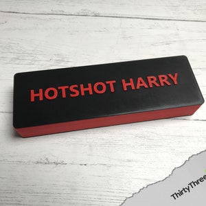 Pode incluir: Uma caixa retangular preta e vermelha com o texto "HOTSHOT HARRY" impresso em vermelho na parte superior.