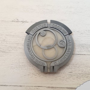 May include: A gray 3D printed circular badge with the text "FOR THE GOOD OF ALL DAUGHTERS NINE TO COMMAND" around the edge. The center of the badge features a stylized design with three concentric circles.