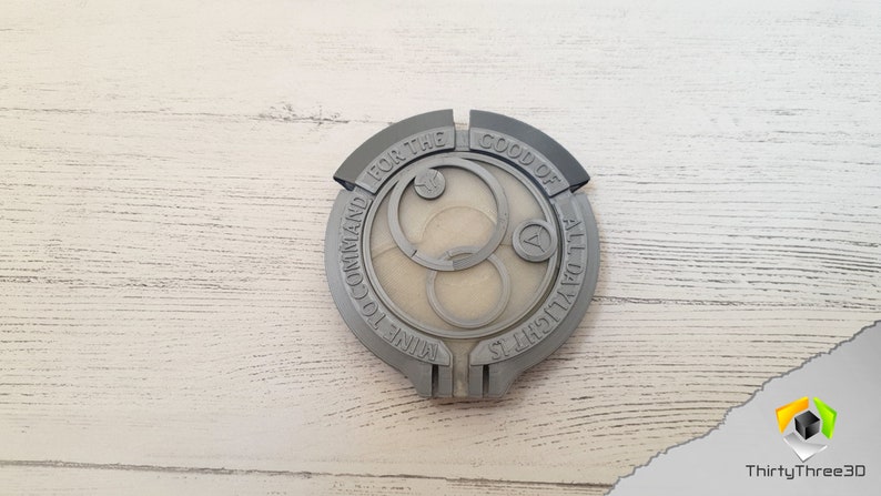 May include: A gray 3D printed circular badge with the text "FOR THE GOOD OF ALL DAUGHTERS NINE TO COMMAND" around the edge. The center of the badge features a stylized design with three concentric circles.