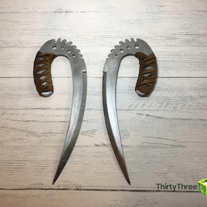May include: Two silver-colored curved blades with brown leather wrapped handles. The blades are sharp and pointed. The blades are on a white wooden background.