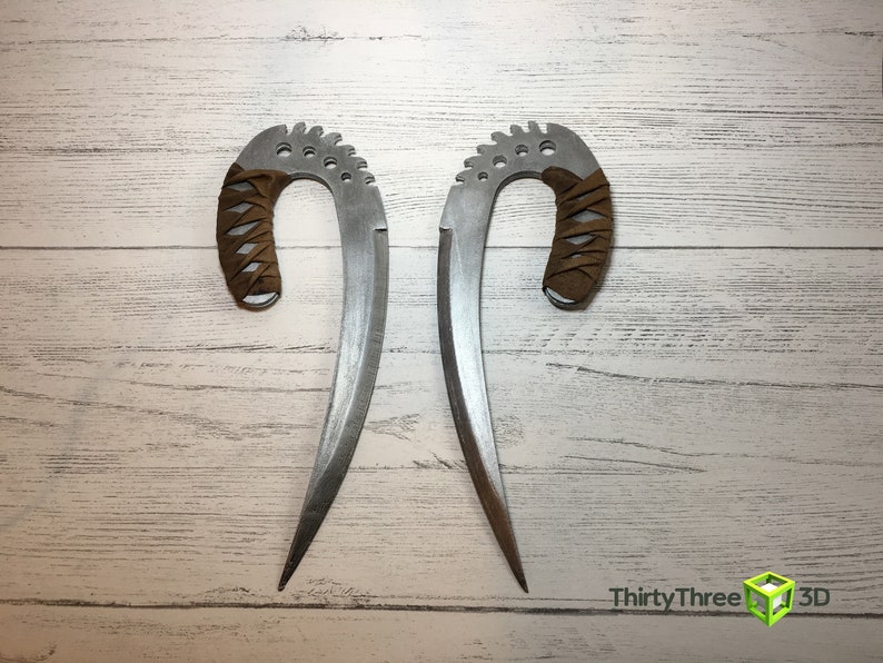 May include: Two silver-colored curved blades with brown leather wrapped handles. The blades are sharp and pointed. The blades are on a white wooden background.