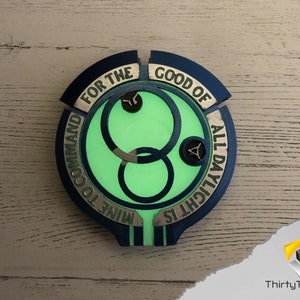 May include: A blue and gold circular badge with a green glow in the center. The badge has the text "FOR THE GOOD OF ALL DAYLIGHT IS MINE TO COMMAND" around the edge.