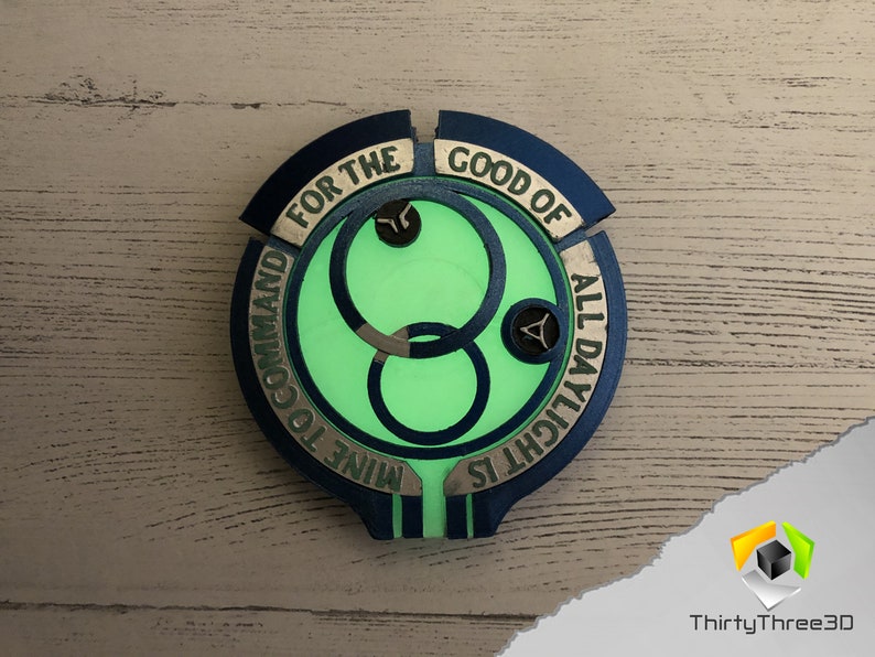 May include: A blue and gold circular badge with a green glow in the center. The badge has the text "FOR THE GOOD OF ALL DAYLIGHT IS MINE TO COMMAND" around the edge.