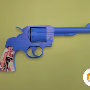 Op de afbeelding: Een blauw speelgoedrevolver met een oranje punt en een handvat met een vintage pin-up girl afbeelding. Het speelgoedpistool staat tegen een lichtgroene achtergrond.