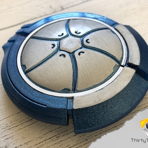 May include: A blue and silver 3D printed disc with a geometric design. The disc has a raised center section with a hexagonal pattern and a textured surface.