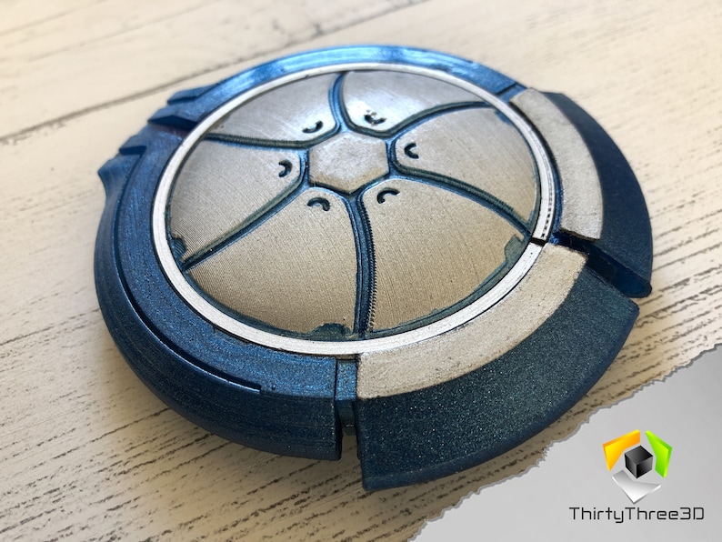 May include: A blue and silver 3D printed disc with a geometric design. The disc has a raised center section with a hexagonal pattern and a textured surface.