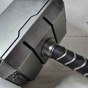 May include: A silver-colored hammer with a black and silver handle. The hammer head has a detailed design and the handle has a textured grip.