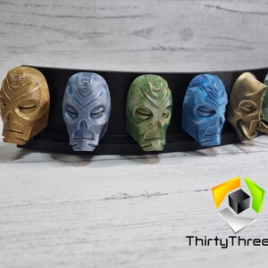 May include: A set of seven 3D printed masks, each with a unique design and color. The masks are arranged in a row on a black display stand. The masks are made of a smooth, plastic-like material and have a variety of colors, including gold, blue, green, and brown.