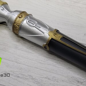 Pode incluir: Um close-up de um cajado decorativo prateado e preto com detalhes dourados. O cajado tem um design detalhado com um s&iacute;mbolo estilizado na parte prateada.