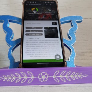Peut inclure: Un support de t&eacute;l&eacute;phone en bois violet avec un motif floral blanc. Le support est en forme de papillon avec des ailes bleues. Un smartphone est dans le support, affichant un site web pour les services d'impression 3D. Le texte du site web comprend "Shop", "Contact us", "Customer photos", et "3D Printing".