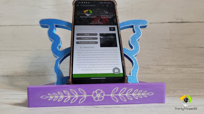 Peut inclure: Un support de t&eacute;l&eacute;phone en bois violet avec un motif floral blanc. Le support est en forme de papillon avec des ailes bleues. Un smartphone est dans le support, affichant un site web pour les services d'impression 3D. Le texte du site web comprend "Shop", "Contact us", "Customer photos", et "3D Printing".