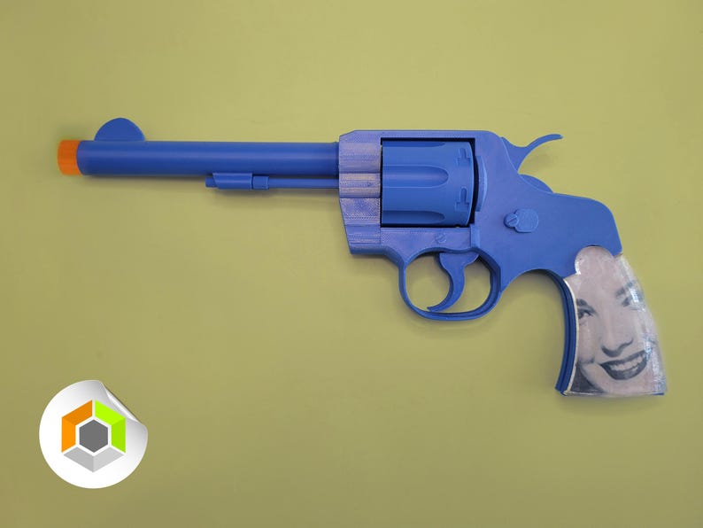 Op de afbeelding: Een blauw speelgoedrevolver met een oranje punt en een lachend gezicht op het handvat. Het speelgoedpistool staat tegen een geelgroene achtergrond. De revolver is een novelty-artikel.