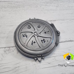 May include: A silver-colored metal disc with a circular design and five sections. Each section has a different symbol inscribed in black. The disc has a raised center with a small hole in the middle.