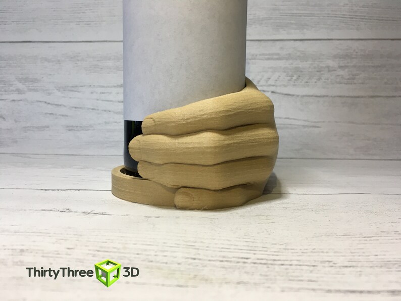 May include: A wooden hand-shaped bottle holder, holding a wine bottle with a white label. The holder is made of light brown wood and has a natural wood finish.