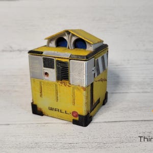 May include: A yellow and gray WALL-E robot toy with a white roof and blue eyes. The robot is made of wood and has a weathered look.