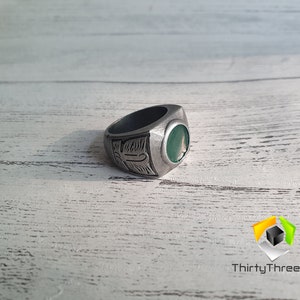 May include: A silver ring with a green stone and an engraved design. The ring is on a white wooden surface.