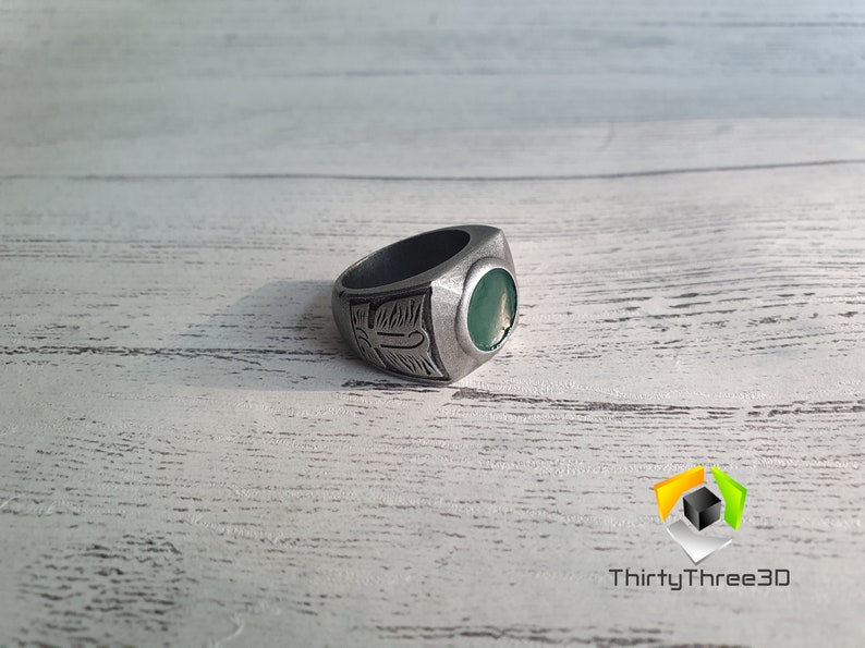 May include: A silver ring with a green stone and an engraved design. The ring is on a white wooden surface.
