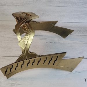 May include: A gold-colored plastic headband with a geometric design. The headband is curved and has a pointed shape on each end.