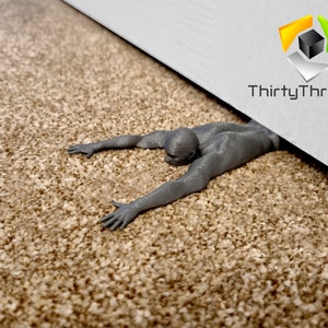 May include: A gray 3D printed doorstop shaped like a person lying on their stomach with their arms outstretched. The doorstop is on a beige carpet. The text "ThirtyThree3D" is visible on a white door.