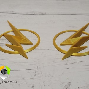 Op de afbeelding: Twee goudkleurige 3D-geprinte Flash-logo's. Elk logo heeft een bliksem in een cirkel.
