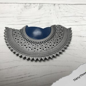 May include: A silver-colored, decorative, half-circle pendant with a blue center. The pendant has a detailed, intricate design with a scalloped edge.