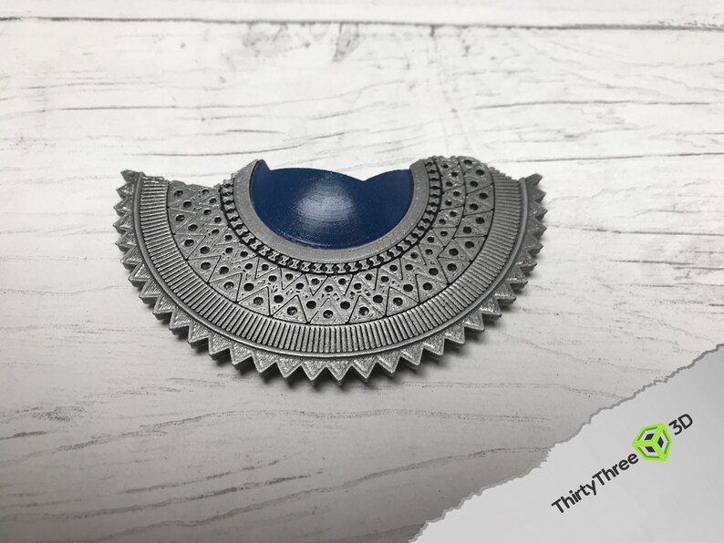 May include: A silver-colored, decorative, half-circle pendant with a blue center. The pendant has a detailed, intricate design with a scalloped edge.
