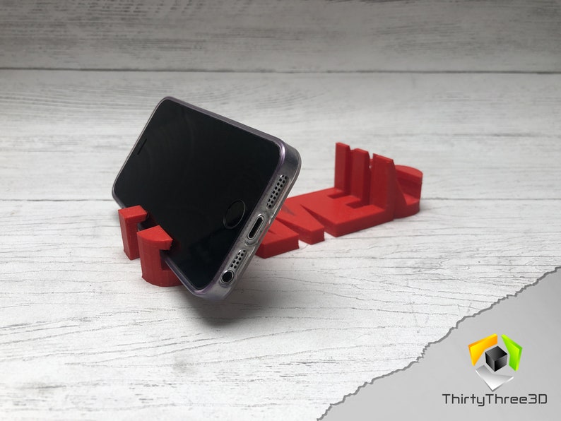 May include: A red 3D printed phone stand shaped like the word "TIME". A black smartphone is resting on the stand.